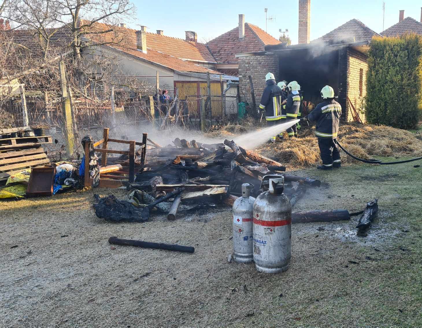 Tűzoltóink teljes védőfelszerelésben, sisakban oltanak egy leégett melléképületet egy udvaron; vízsugár a parázsló farakásra.